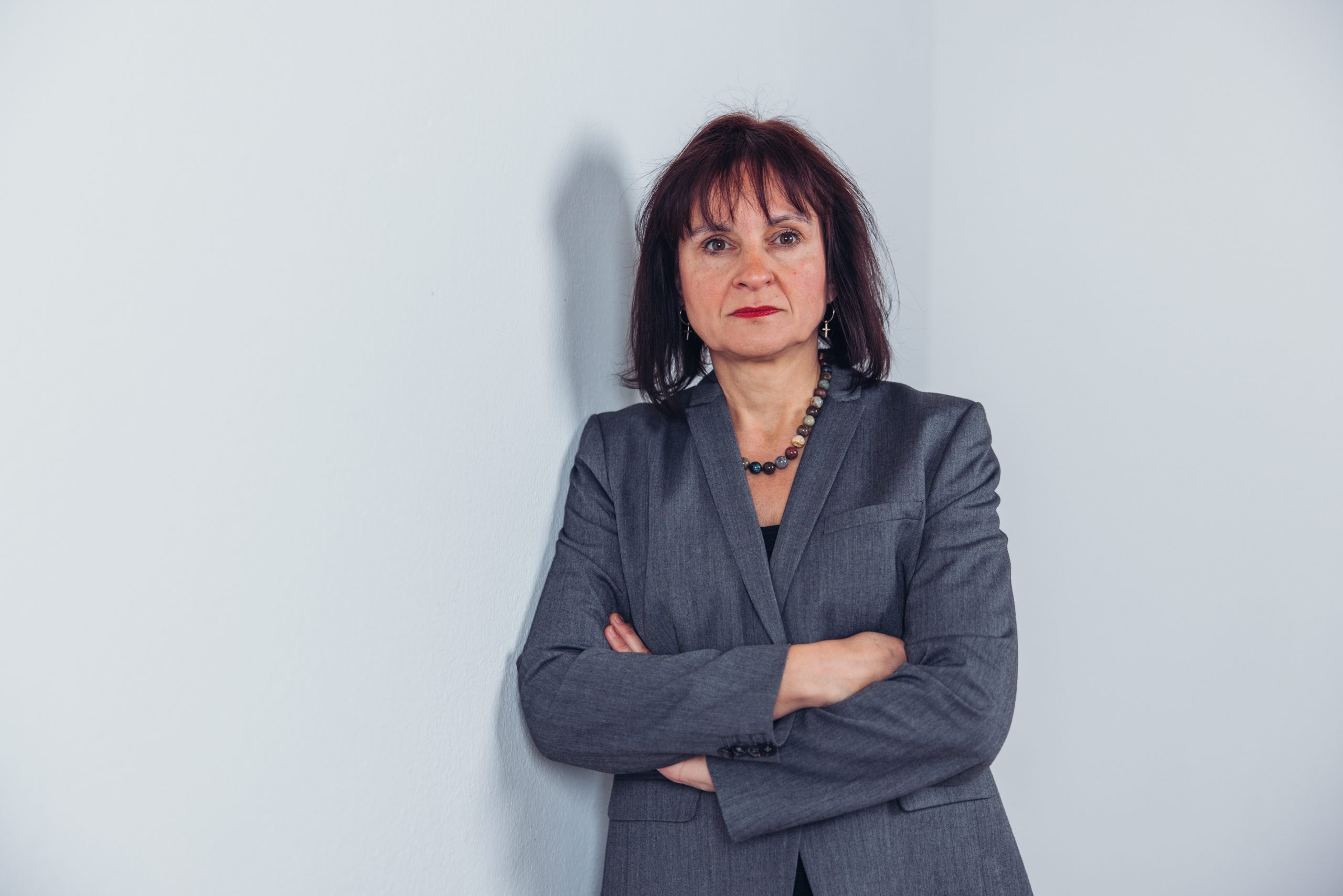 Frau mit dunklen Haaren und grauem Anzug steht vor weißer Wand mit verschränkten Armen.