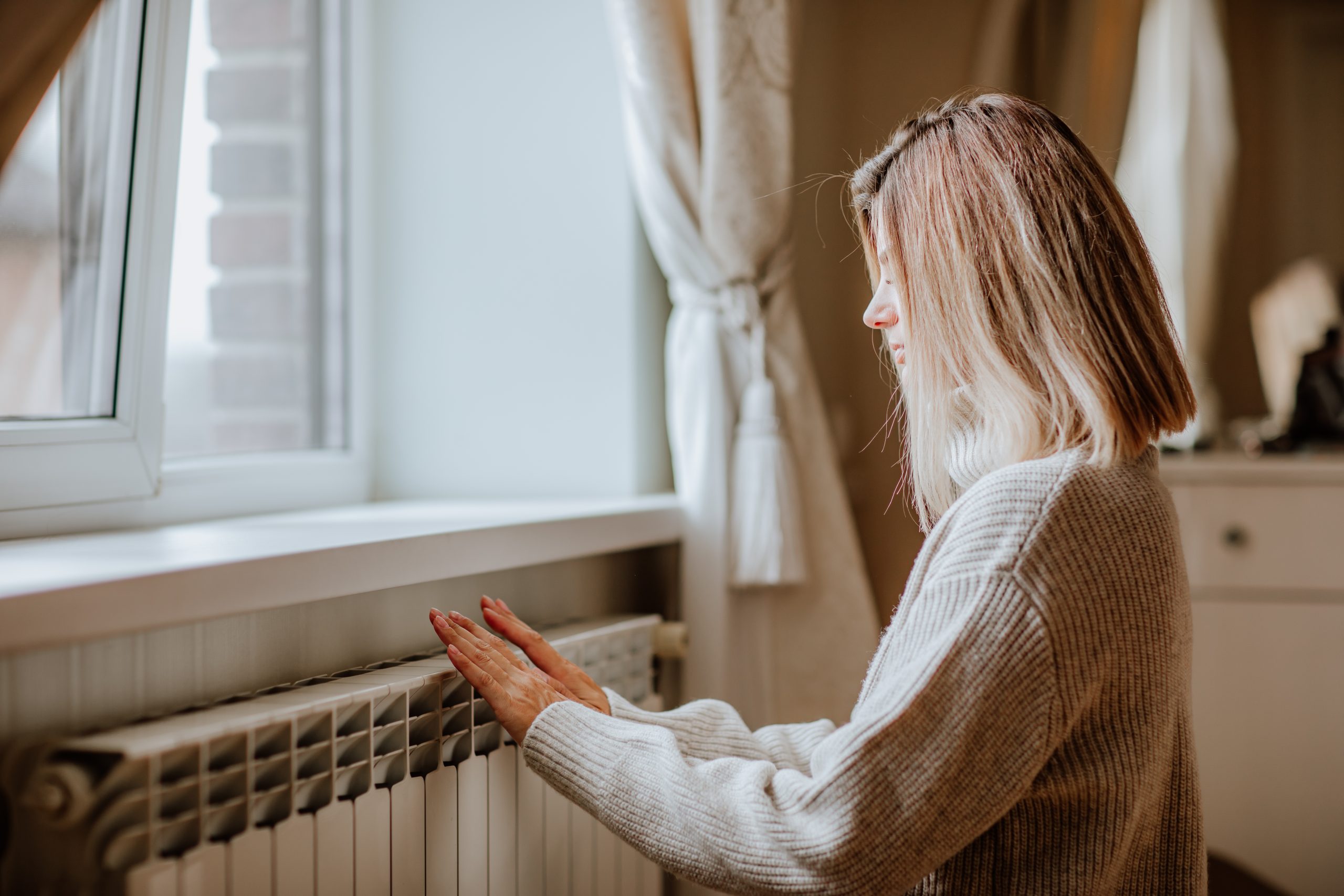 Young blond woman in long winter beige sweater is posing at home near the radiator. Winter season concept. Economy program